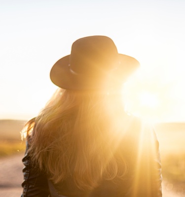 A woman in a hat and leather jacket strolls along a road, illuminated by the warm hues of a sunset.